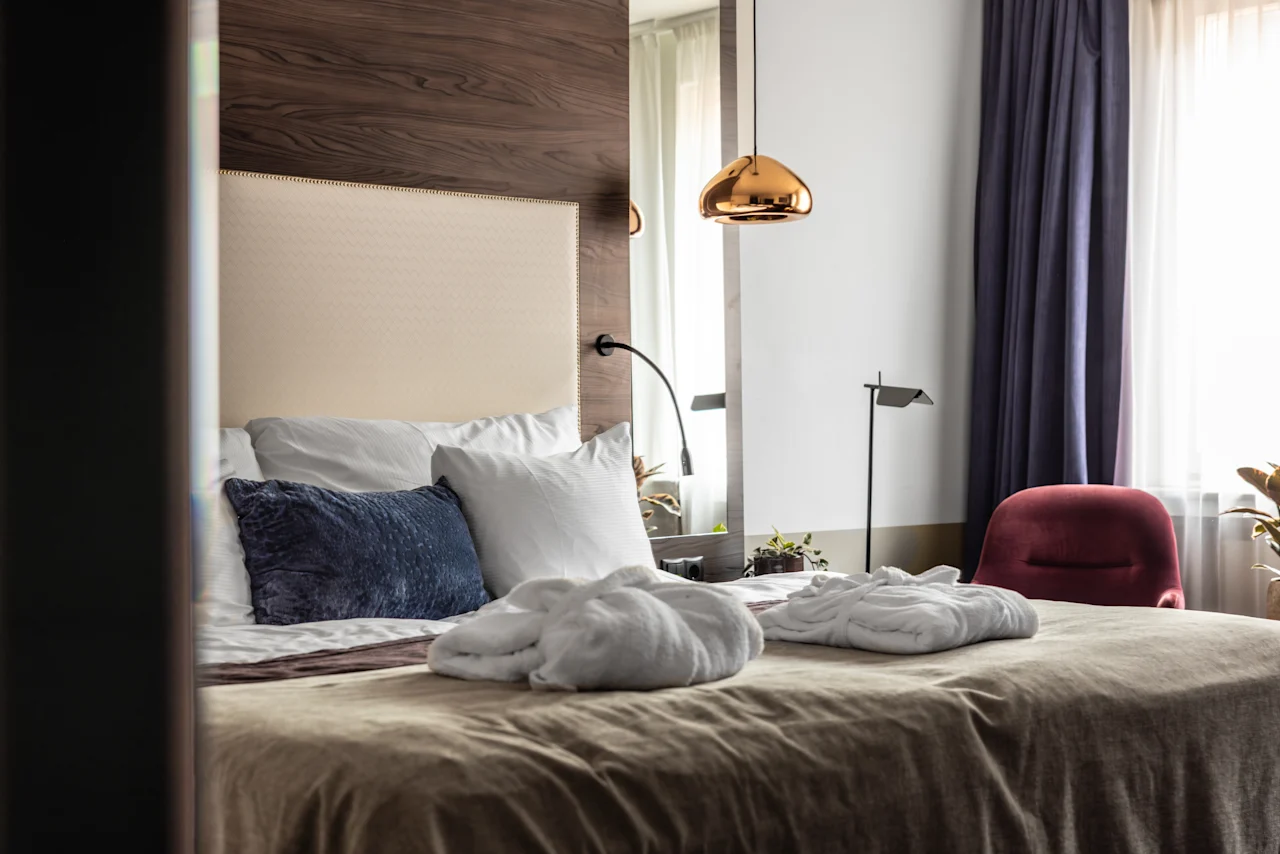 Kodikas hotellihuone, jossa sänky on siististi pedattu tyynyillä ja kylpytakeilla. Pehmeä valo suodattuu verhojen läpi tyylikkään punaisen tuolin vieressä. Lämmin, kutsuva tunnelma täyttää tilan.