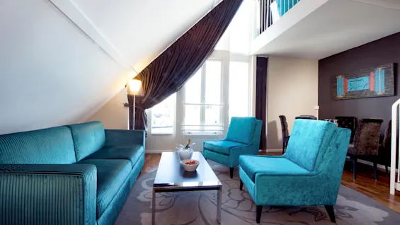 A teal sofa and chairs furnish a modern living room with a chrome coffee table, under a slanted ceiling with draped curtains.
