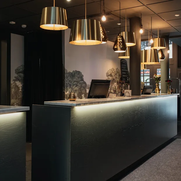 Et indbydende hotelreceptionsområde med en moderne disk med varm belysning, messingpendellamper og et naturinspireret vægmaleri.