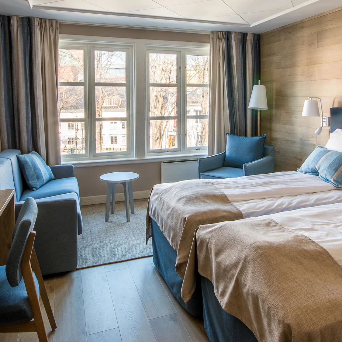 Et hotellrom med to pent redes senger og et skrivebord i tre; naturlig lys strømmer inn gjennom store vinduer og viser en byscene utenfor.