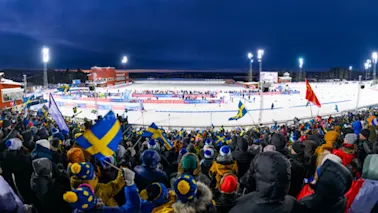 En stor folkemengde, mange viftende med flagg, ser på et vintersportsarrangement på en opplyst, snødekt bane om natten. Banner: BAUHAUS.