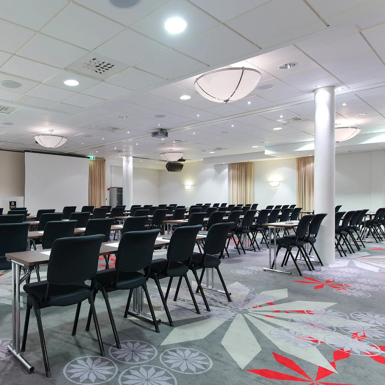 Rows of black chairs and tables are arranged in a conference room with a projector and patterned carpet.