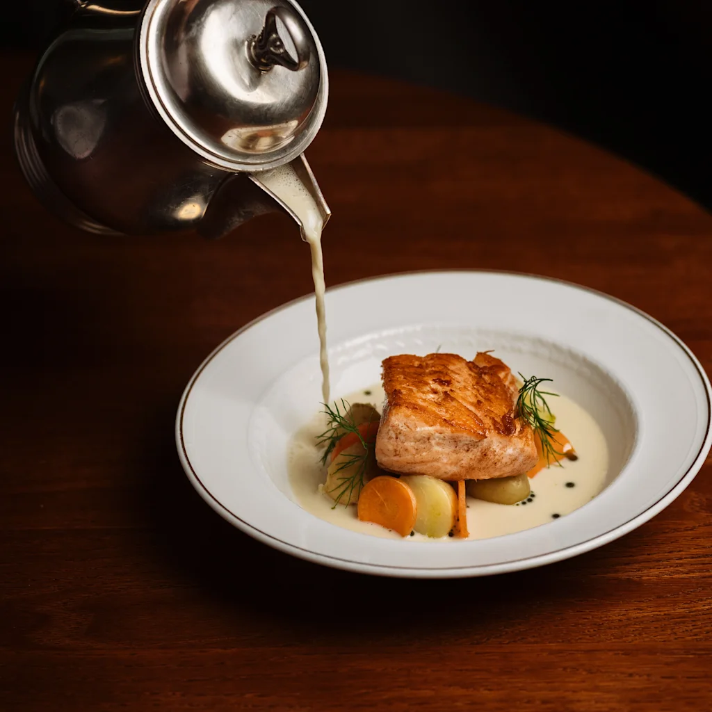 En elegant rett med laks og grønnsaker får en siste touch når en kremet saus helles fra en sølvkanne.