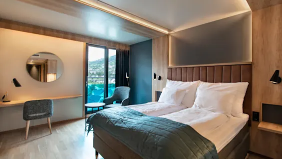 En prydligt bäddad säng med vita kuddar och ett blått täcke står i ett modernt hotellrum. Rummet har ett skrivbord, stol, rund spegel och utsikt genom glasdörrar.