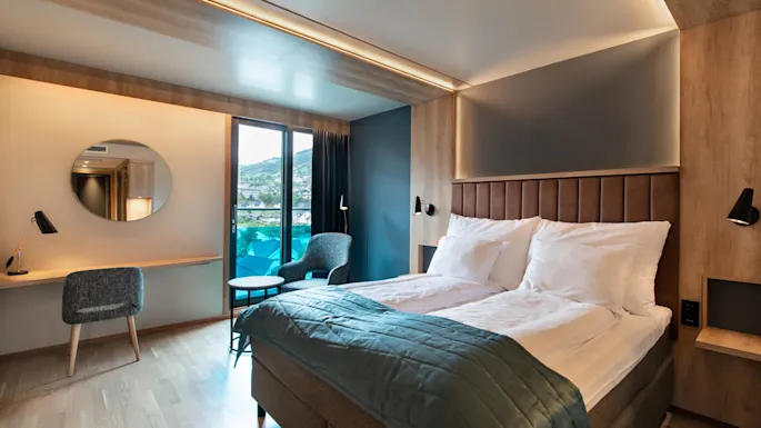 A neatly made bed with white pillows and a blue quilt sits in a modern hotel room. The room features a desk, chair, circular mirror, and a view through glass doors.