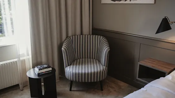 Ett mysigt rumshörn med en randig fåtölj, ett litet bord med böcker och ett fönster med ljusa gardiner, perfekt för avkoppling.