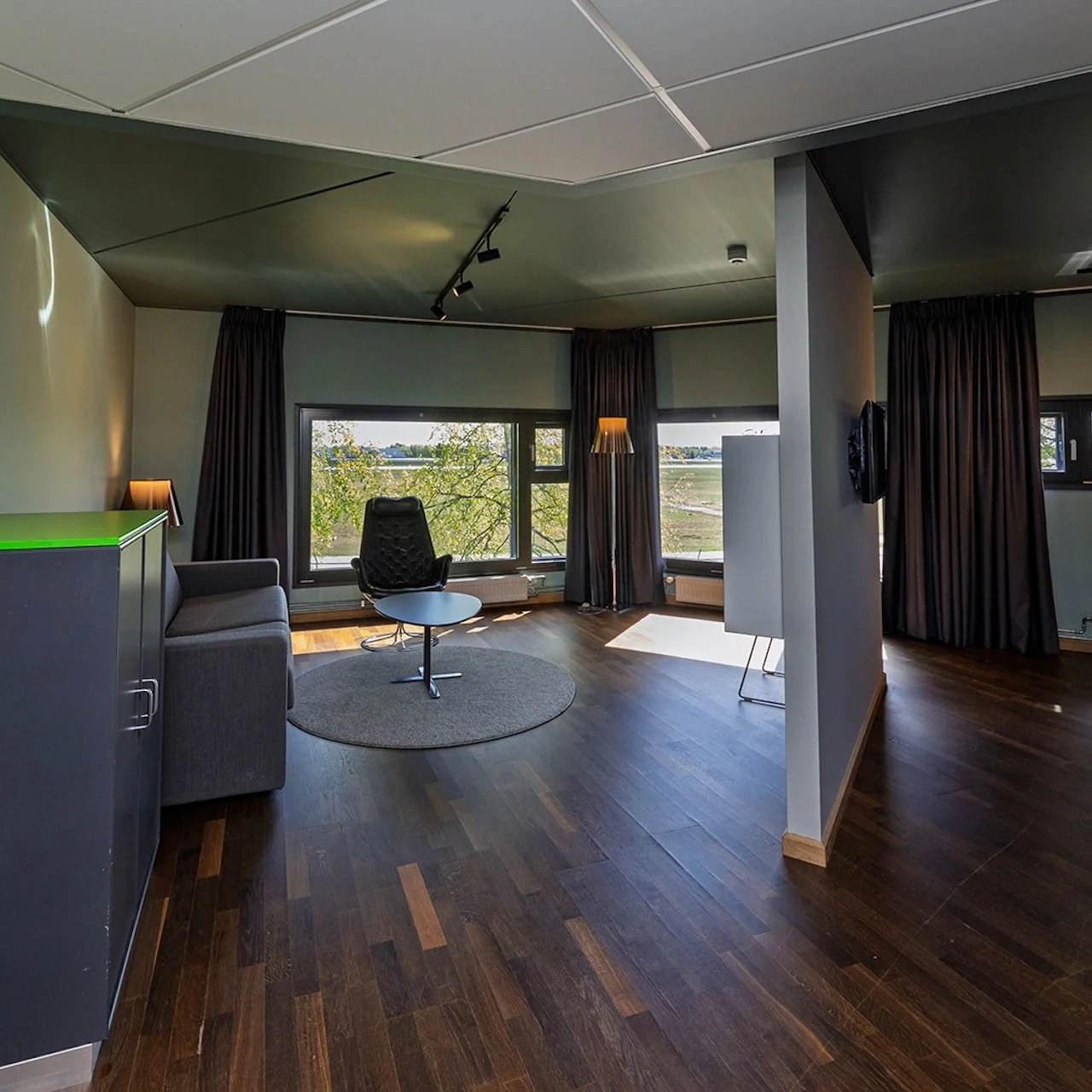 A modern room with a black swivel chair on a rug, facing large windows. A gray sofa, bed, cabinets, and curtains complement the space, illuminated by lamps.