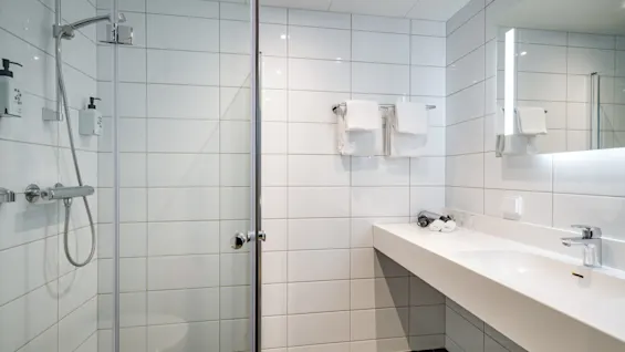 Et moderne hotellbad med dusj, en stor servant med vask og et speil. Såpedispensere leser HAIR & BODY.