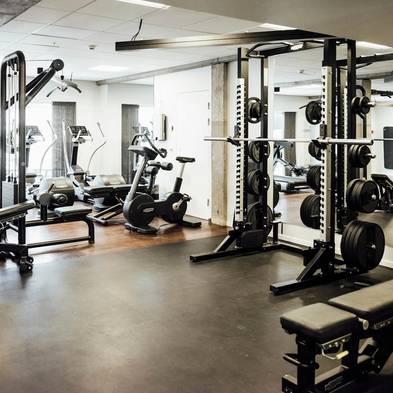 Træningsudstyr i brug, med løbebånd, motionscykler og vægtløftningsstationer i et organiseret, veloplyst fitnesscenter med trægulv og spejle på væggene.