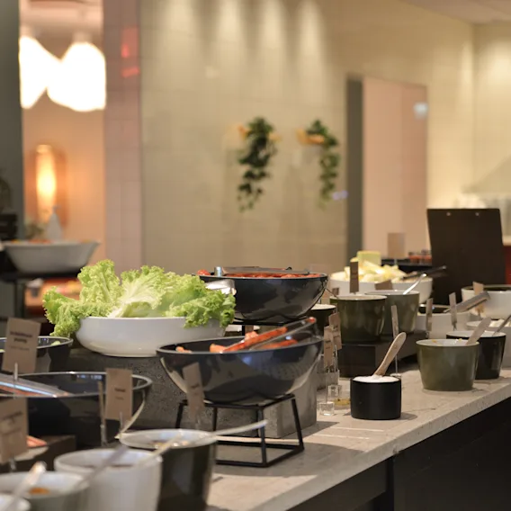 A buffet table displays various salads in bowls. Guests serve themselves amid warm lighting and soft decor, with serving utensils readily available for convenience.