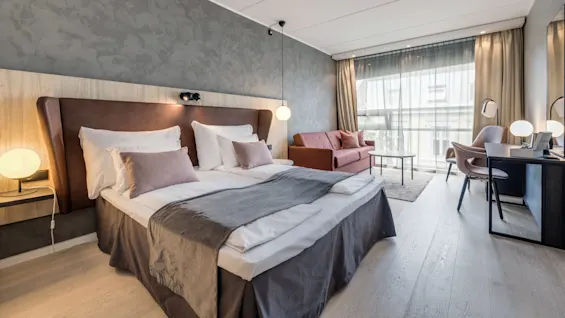 A neatly made double bed with plush pillows and a dark bedspread stands in a modern hotel room. Nearby, a sofa, desk, and large window provide a spacious, bright atmosphere.