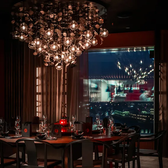Elegant lysekrone henger over et trebord dekket med glass og dekorasjoner i en dempet, stilig restaurant. Et stort vindu avslører en byutsikt utenfor.