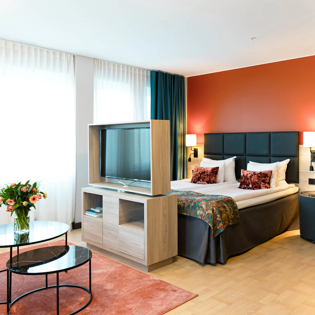Ett modernt hotellrum har en prydligt bäddad säng med kuddar, sänglampor, en soffstol, en TV på en träställning och runda soffbord, mot eleganta gardiner.