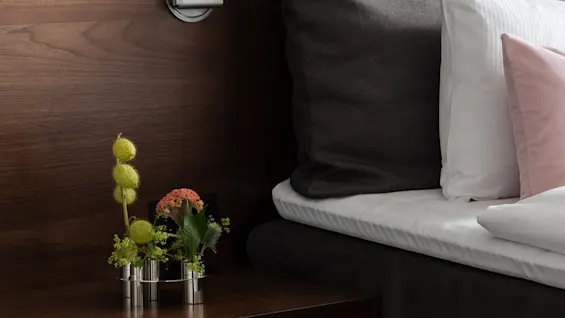 Viihtyisä sängynvierusalue Stratum Suitessa Clarion Hotel Signissa Tukholmassa, jossa on puinen yöpöytä kukkineen ja sänky tyynyineen.