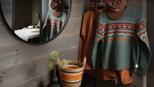 A round mirror on a wooden wall reflects a colorful sweater and cap hanging on a black coat rack. Below, a black table holds a vase and a woven basket. Text: "Please make up room".