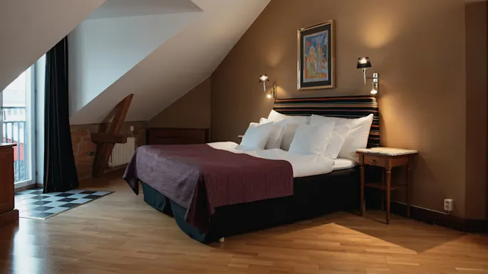 En stor seng med flere puter og et lilla overkast står under et skråtak. To vegglamper lyser opp et maleri, mens tremøbler og murverk skaper en koselig atmosfære.