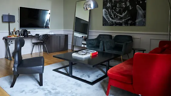 Black coffee table holds books, surrounded by red and black sofas and chairs. Large television and mirror adorn the wall, with a side table and decor items adjacent.