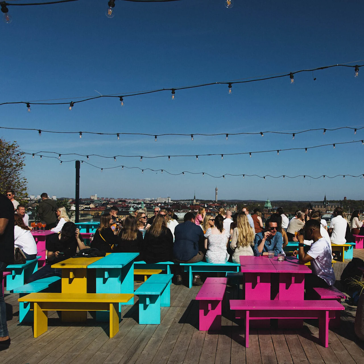 Fargerike benker omgir grupper av mennesker som nyter det solrike været på en takterrasse, med lyslenker over hodet og en byutsikt i bakgrunnen.