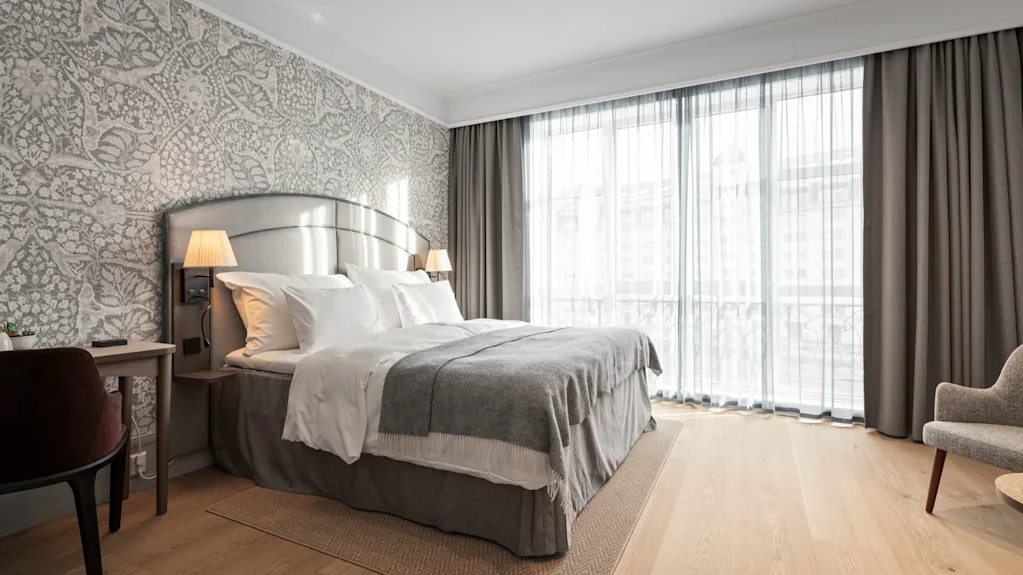 A bright and inviting hotel room with a large bed, elegant patterned wallpaper, and sheer curtains letting in natural light.