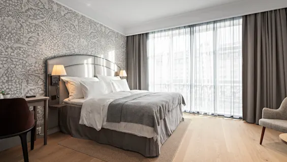 A bright and inviting hotel room with a large bed, elegant patterned wallpaper, and sheer curtains letting in natural light.