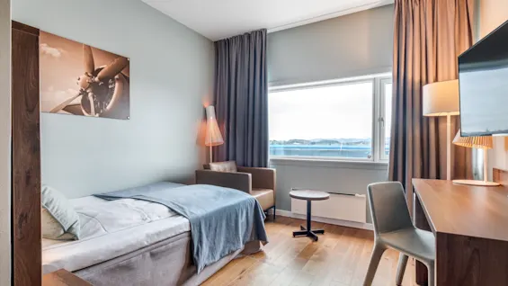 A tidy hotel room with a single bed and blue blanket, wooden desk, chair, and standing lamp. Large window with drapes shows an outdoor view. Wall art depicts a vintage airplane propeller.