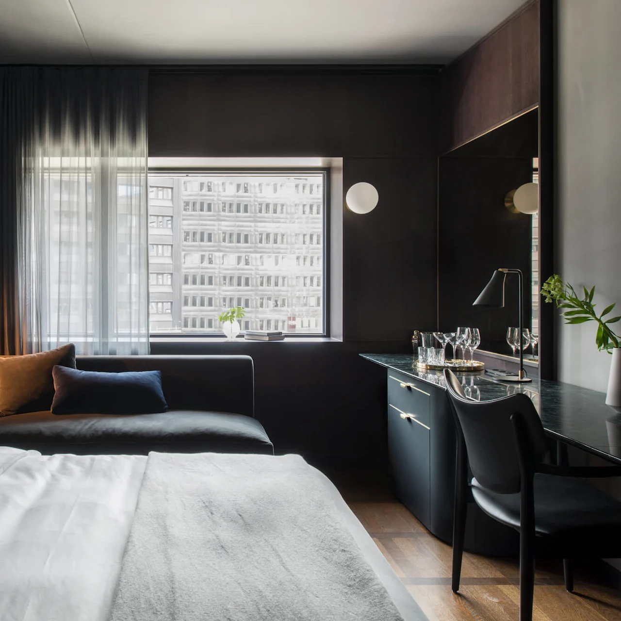 En pent senger står i et moderne hotellrom med tregulv. En sofa står nær et vindu med gjennomskinnelige gardiner og et skrivebord med glass og lampe.
