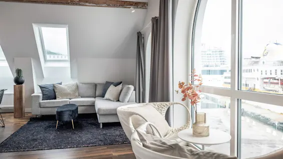 Beige soffa i ett modernt vardagsrum bredvid stora välvda fönster. Ett litet bord med en blomanordning och två runda stolar förstärker den mysiga atmosfären.