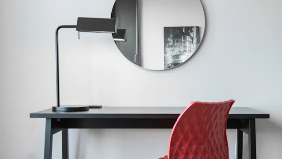 A black desk lamp on a black table with a red chair and a round mirror reflecting a monochrome wall art piece in a minimalist room.