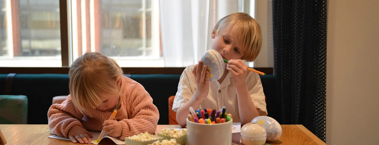 Två barn ritar och färglägger vid ett träbord fullt med pennor, böcker och popcorn under ett stort fönster.