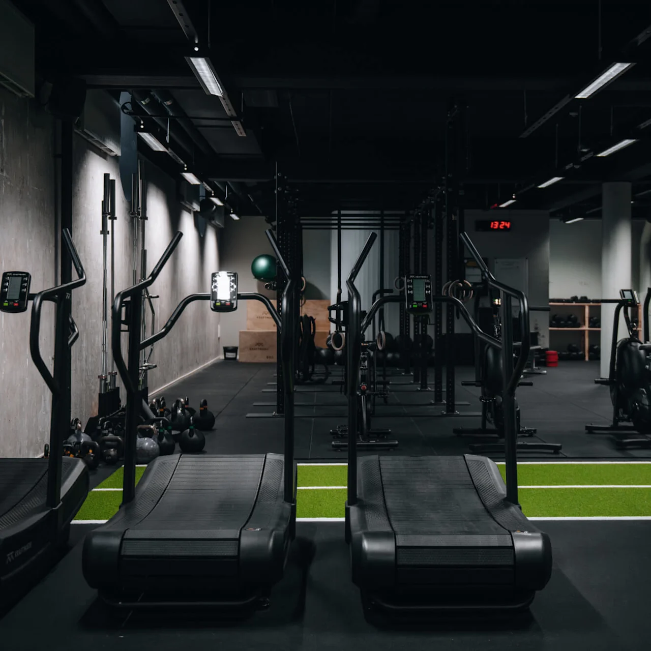 Löpband står stilla i ett svagt upplyst gym med svart golv och en timer som visar "13:24" på väggen ovanför olika gymutrustningar.