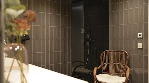 En kurvestol med en pude står stille i et flisebelagt badeværelse ved siden af en bruser med mørke glasdøre og en bordplade med en vase med blomster.