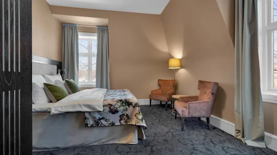 Elegant inrett hotellrum med blommigt överkast, två fåtöljer och mjuk belysning från ett fönster.