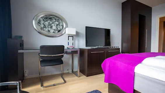 Et moderne rom har en svart stol ved et glassbord under et ovalt kunstverk. En TV står på et treskap ved siden av en seng med et lilla teppe.