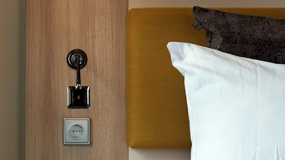 En seng med hvide puder og et tæppe er ved siden af et træpanel. En lampe og stikkontakt er over en lille hylde med en grøn hæfte.