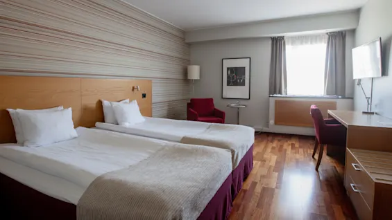 Two neatly made twin beds stand beside a striped wall. A red chair and small table are near a window with curtains. A desk and TV complete the room's simple furnishings.