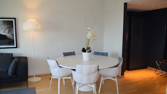 A white round table is surrounded by six chairs, topped with a vase of flowers. Adjacent is a standing lamp and a dark sofa, set on a wooden floor.