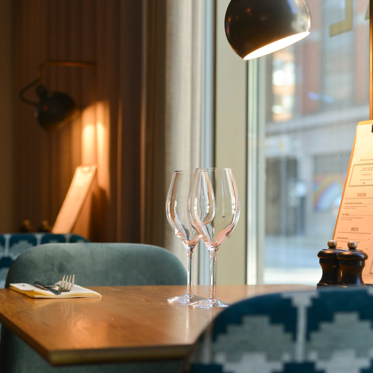 Et træbord i en restaurant har tre tomme vinglas og en menu mærket "SOCIAL," under en moderne lampe, nær et stort vindue med byudsigt.