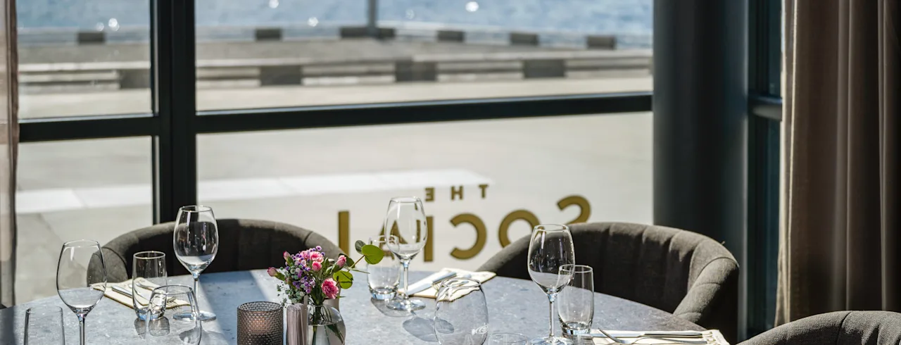 Et dækket spisebord med glas og bestik, med udsigt over en naturskøn havnefront med grønne bakker. Tekst på vindue: THE SOCIAL.