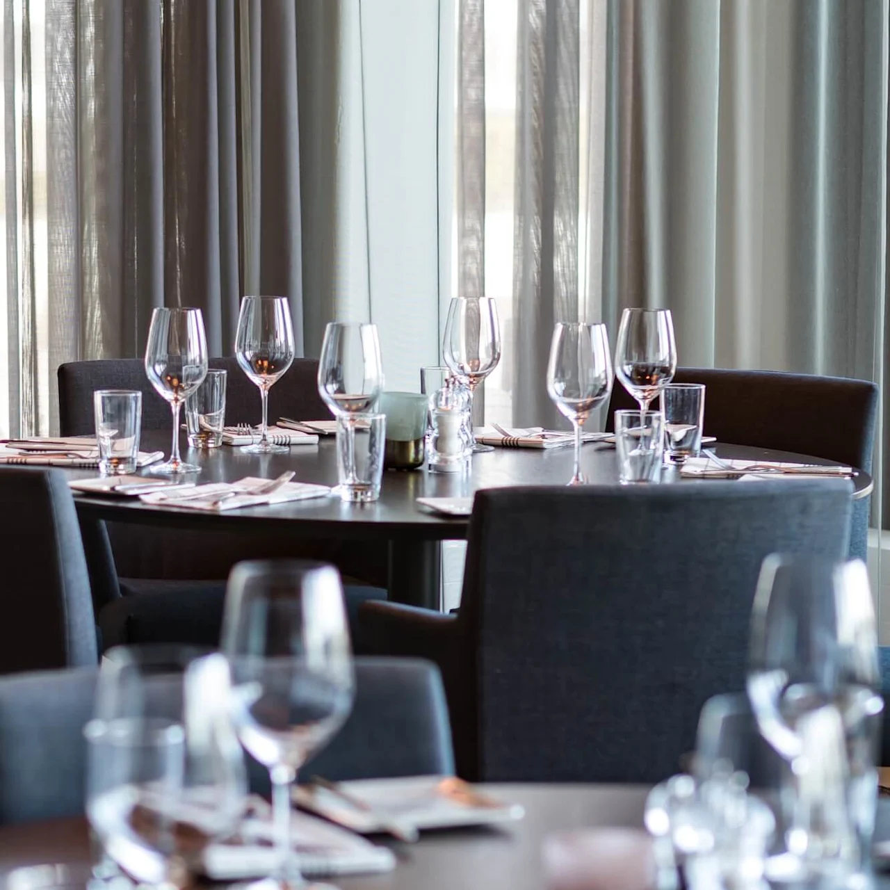 A round table set with wine glasses and napkins, surrounded by upholstered chairs in a softly lit dining area with sheer curtains.
