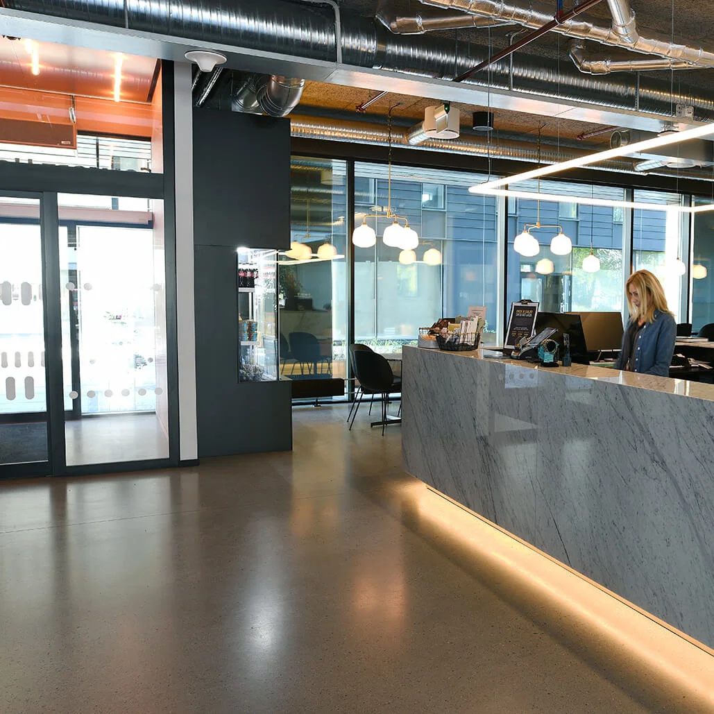 Reception med en kvinde, der arbejder, omgivet af moderne indretning og hængende lamper. Et tv på væggen viser en nyhedsoplæser. Glasdøre fører til et siddeområde.