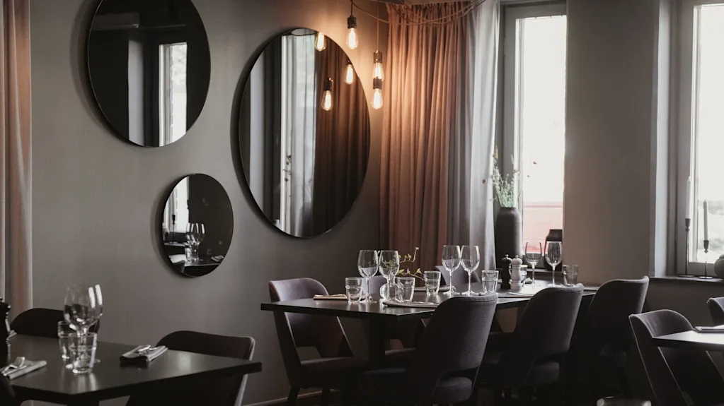 Tables set with wine glasses and cutlery in a dimly lit restaurant. Mirrors adorn the gray walls, reflecting warm hanging lights. Soft curtains and windows enhance the cozy atmosphere.
