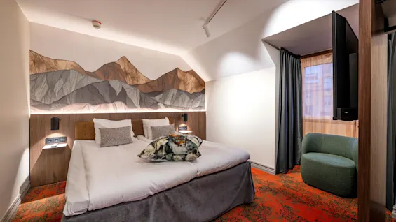A bed with white linens and decorative pillows is centered beneath a mountain-themed mural. The room has patterned red carpet and a green chair by a window with sheer curtains.