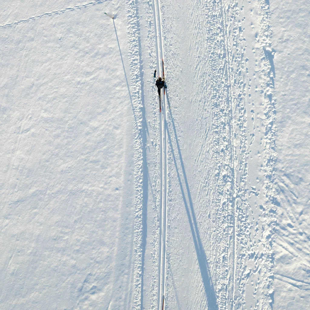 Ilmakuva kahdesta ihmisestä hiihtämässä hoidetulla ladulla laajassa lumisessa maisemassa, heittäen pitkiä varjoja.
