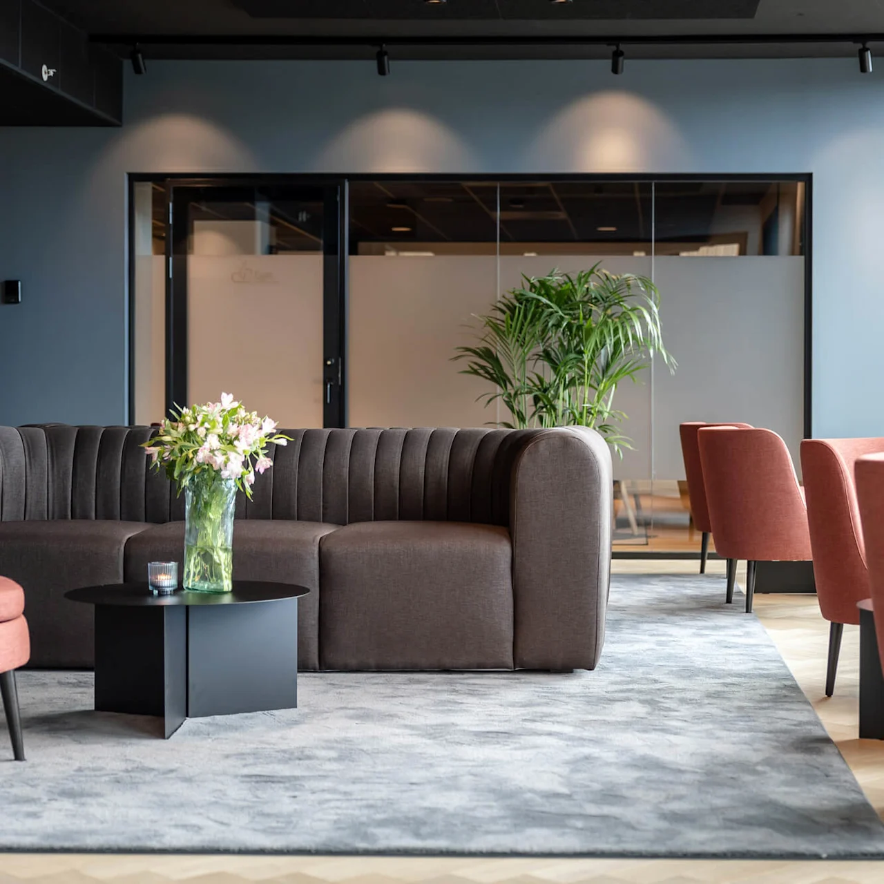 En brun sofa står på et gråt tæppe, som holder en vase med blomster på et sort bord. Omgivet af lyserøde stole, store vinduer og en grøn plante i nærheden.