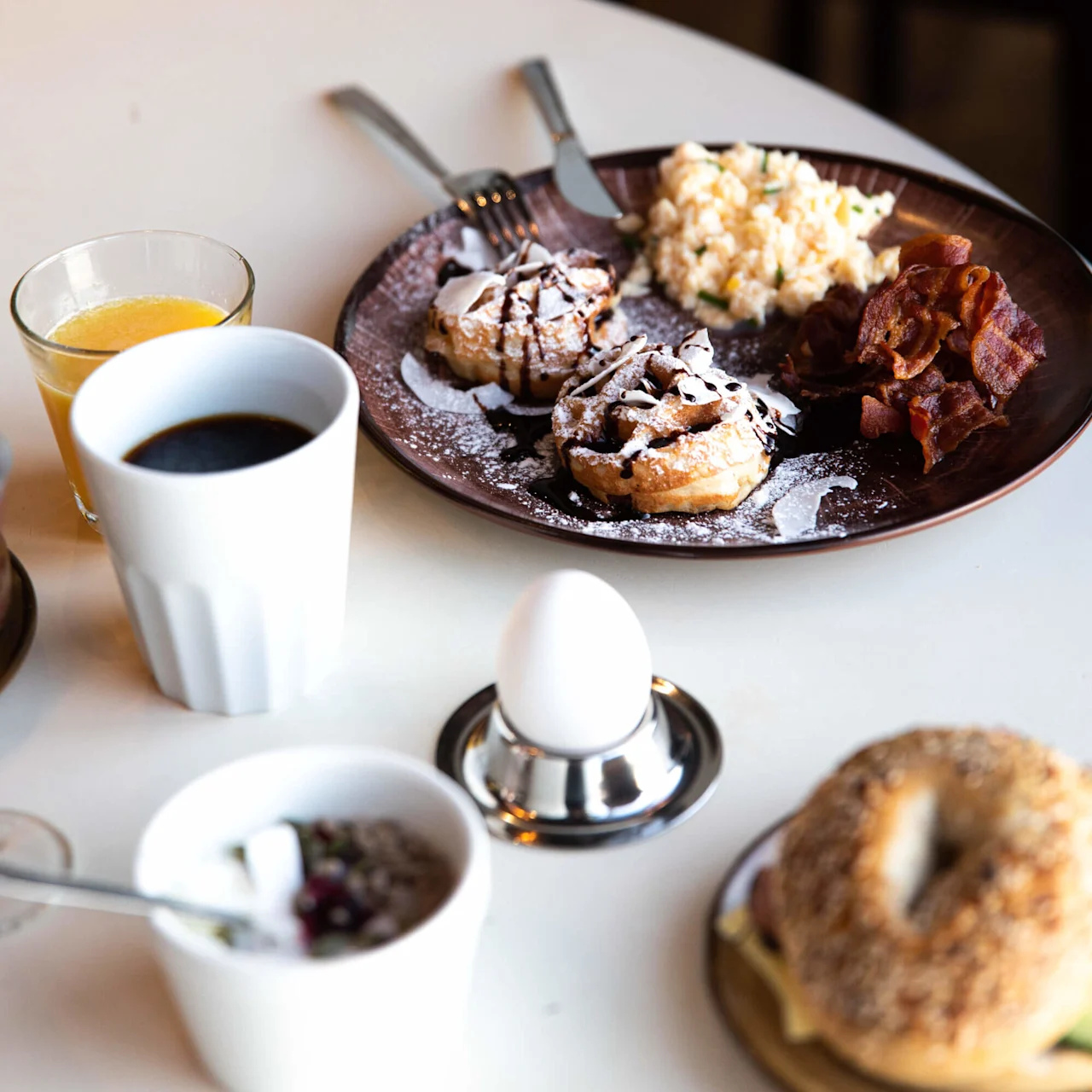 Pancakes drizzled with chocolate, scrambled eggs, and crispy bacon sit on a plate on a table. Nearby are a cup of coffee, orange juice, a boiled egg, and a bagel with cream cheese.