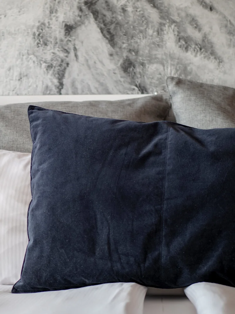 En marinblå kudde vilar mot två större grå kuddar på en vit säng med en grå texturerad vägg i bakgrunden.