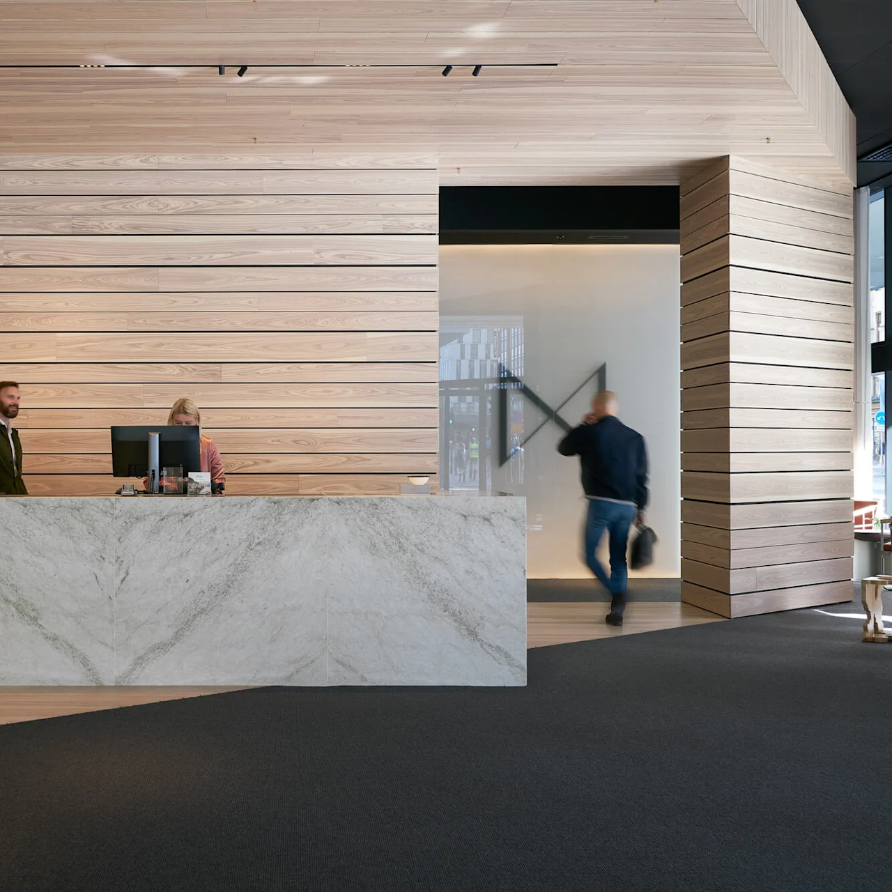 Reception desk with two people standing behind computers, interacting. Modern wooden panel wall background, marble desk. Another person walks by carrying a bag. Bright, spacious interior.