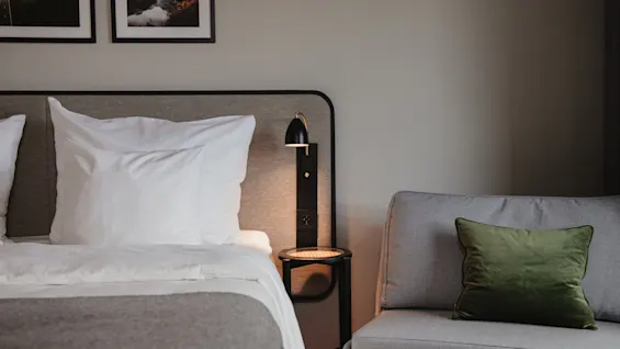 Seng med hvite puter og grått teppe, ved siden av et lite sidebord og lampe, ved siden av en grå sofa med en grønn pute i et mykt belyst soverom.
