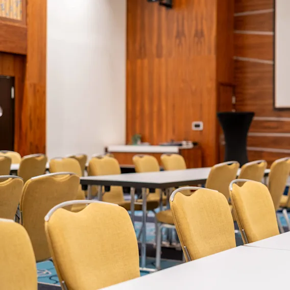 Rader med gule stoler er vendt mot en stor hvit projektorskjerm i et trepanelert konferanserom, med bord arrangert for et møte eller en presentasjon.