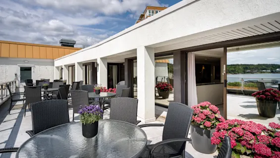 En innbydende utendørsterrasse med bord, stoler og fargerike potteplanter, som tilbyr en naturskjønn utsikt over vannet under en delvis skyet himmel.